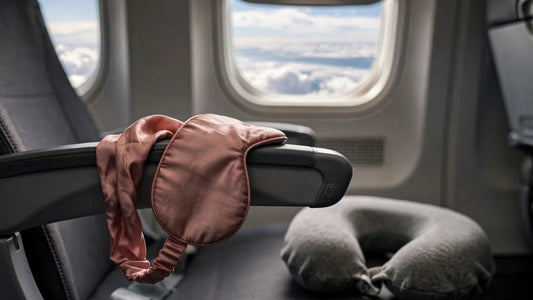 Masque de sommeil en soie rose poudré posé sur l'accoudoir d'un siège d'avion près du hublot