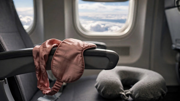 Masque de sommeil en soie rose poudré posé sur l'accoudoir d'un siège d'avion près du hublot